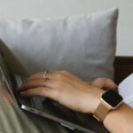 Smart Home Integration - Side view of crop unrecognizable female freelancer in casual clothes and smartwatch lying on comfortable sofa and working remotely on laptop