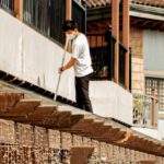 Roof Maintenance - Male cleaner with broom wearing mask sweeping roof of small residential building with outdoor cafeteria on sunny weather
